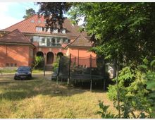 Ein zweistöckiges Gebäude mit roter Fassade und einem steilen Ziegeldach, umgeben von Bäumen und einem Garten. Im Vordergrund sind ein Trampolin, geparkte Autos und ein gepflegter Rasen zu sehen.