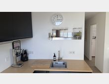 Moderne Küchenzeile mit Holzarbeitsplatte und eingelassenem Edelstahlspülbecken, darüber eine Wanduhr und ein Metallregal mit Vorratsgläsern und Obstkorb. Links sind dunkle Hängeschränke, eine Kaffeemaschine und ein Messerblock zu sehen; rechts führt ein Durchgang in einen hellen Flur.