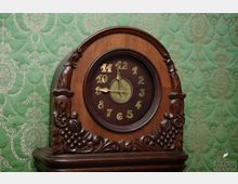 Holzuhr mit geschnitzten Verzierungen, darunter Frucht- und Blattmotive, vor einer grünen, mit goldenem Ornamentmuster versehenen Tapete. Die Ziffern der Uhr sind goldfarben, und das Zifferblatt ist dunkel.