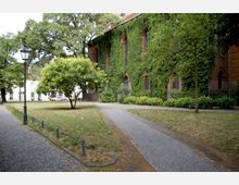 Grüne Parklandschaft mit gepflasterten Wegen, einer Laterne und einem Baum im Vordergrund. Im Hintergrund ist ein mit Efeu bewachsenes Gebäude aus rotem Backstein mit gotischen Fenstern zu sehen.