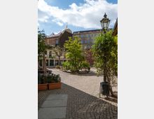 Ein gepflasterter Innenhof mit Bäumen und Sitzbänken, flankiert von Gebäuden im historischen Stil. Im Hintergrund ist eine goldene Kuppel sichtbar, die zu einer Synagoge oder einem ähnlichen Bauwerk gehört, während eine alte Straßenlaterne und Pflanzkübel im Vordergrund stehen.