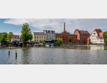 Uferansicht eines Flusses mit mehreren Gebäuden im Hintergrund, darunter rote Backsteinbauten mit Schornstein und Häuser mit hellen Fassaden. Am Ufer sind kleine Boote und Holzhütten zu sehen, während umgebende Bäume die Szene umrahmen.