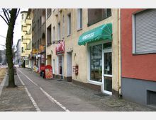 Straßenszene mit mehrstöckigen Wohnhäusern und kleinen Geschäften im Erdgeschoss, darunter ein Hundesalon mit grünem Vordach. Der Gehweg aus Kopfsteinpflaster und ein markierter Fahrradweg verlaufen parallel zur Straße, auf der linken Seite steht eine Baumreihe.