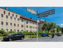 Straßenkreuzung in einem Wohngebiet mit beige verputztem Mehrfamilienhaus im Hintergrund, dekoriert mit Wandreliefs und Blumenkästen an einigen Fenstern. Im Vordergrund sind Straßenschilder der "Winfriedstraße" und "Charlottenburger Straße" sowie parkende Autos und ein Rollerfahrer zu sehen, umgeben von Grünflächen und Bäumen.