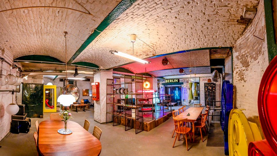 Großer, loftartiger Innenraum mit weiß gestrichenen Ziegelgewölbedecken, Sichtinstallationen und industrieller Beleuchtung. Lange Holztische mit Stühlen stehen vor einem abgetrennten Bühnenbereich aus Metallgittern; im Hintergrund sind farbige Neonlichter und ein Schild mit der Aufschrift „BERLIN“ zu sehen.