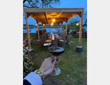 Hand mit Weinglas im Vordergrund, dahinter eine überdachte Sitzplatzfläche im Freien mit Holzpergola, warmen Lichterketten und Blick auf einen Fluss bei Abenddämmerung.