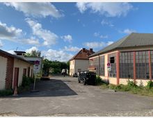 Industriegebiet mit älteren Gebäuden, darunter eine große Halle mit Glasfenstern und ein kleineres Gebäude mit roten Türen. Im Vordergrund ein Parkplatz mit mehreren Fahrzeugen und Verbotsschildern, umgeben von Grünflächen und teilweise bewachsenen Rändern.