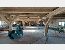 Innenraum eines historischen Fachwerkgebäudes mit Holzbalken und Holzboden. Der Raum zeigt verschiedene landwirtschaftliche Maschinen und Ausstellungsstücke, mit gemauerten Wänden und Fenstern, durch die Tageslicht einfällt.