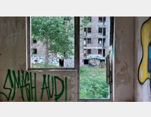 Blick durch ein zerbrochenes Fenster in einem verlassenen Gebäude auf eine verwilderte Grünfläche und ein weiteres verfallenes, mit Graffiti bedecktes Wohngebäude. Die Wände im Vordergrund sind ebenfalls mit Graffiti besprüht.