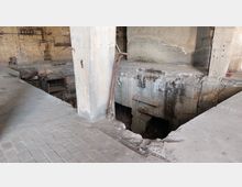 Innenansicht eines verlassenen Industriegebäudes mit beschädigten Betonböden und freiliegenden Stahlträgern. Im Hintergrund sind eine gemauerte Wand, alte Rohre und ein offener Schacht sichtbar.
