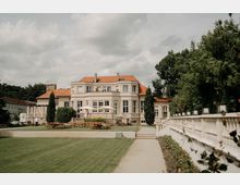 Blick auf eine große, helle Villa mit rotem Ziegeldach und vielen Fenstern, davor eine Terrasse mit Sonnenschirmen und Sitzgelegenheiten. Im Vordergrund führen ein breiter Kiesweg und eine weiße Balustrade durch einen gepflegten Garten mit Rasenflächen und Bäumen.
