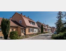 Reihenhaussiedlung mit hell gestrichenen Häusern und roten Satteldächern, flankiert von schmalen Gehwegen, einzelnen Bäumen und Sträuchern. Im Vordergrund eine schmale, asphaltierte Straße mit parkenden Autos unter klarem, blauem Himmel.