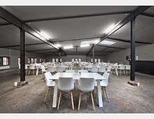 Großer, offener Veranstaltungsraum mit modernem, industriellen Design, erkennbar durch schwarze Metallstützen und eine Betonbodenfläche. Der Raum ist mit weißen Tischen und Stühlen in schlichter Optik eingerichtet; auf den Tischen stehen kleine Blumenvasen.