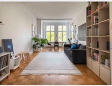 Helles Wohnzimmer mit Holzboden, einem großen grauen Sofa, einem flachen weißen Teppich und einer Bücherwand rechts. Im Hintergrund befindet sich ein Esstisch vor großen Fenstern, die viel Tageslicht hereinlassen; links stehen Pflanzen und kleine Möbel.