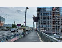 Straßenansicht mit einer Baustelle auf der rechten Seite, wo ein Hochhaus von einem Gerüst umgeben ist. Links befindet sich eine mehrspurige Straße mit Autos und Fußgängern, sowie ein mit Graffiti bedecktes Brückengeländer im Vordergrund.