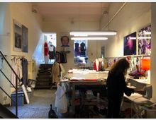 Ein Atelierraum mit offenem Arbeitsbereich. Im Vordergrund steht eine Person, die Kleidung bügelt, während im Hintergrund ein großer Arbeitstisch mit Stoffen und Nähzubehör sowie an der Wand hängende Mode und Poster zu sehen sind. Treppen im hinteren Bereich führen zu einem kleinen Ausstellungsraum.