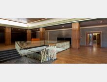 Innenansicht eines Gebäudes im Retro-Stil, mit Holzvertäfelungen, gelb-grünem Geländer um ein Treppenhaus und Parkettboden. Im Hintergrund sind Wände mit grauer Textur, eine hölzerne Doppeltür und mehrere Stützsäulen sichtbar.