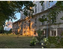 Historisches Gebäude mit verputzter Fassade und rechteckigen Fenstern, umgeben von einem Garten mit Palmen und blühenden Sträuchern. Die Szenerie ist im warmen Licht des Sonnenuntergangs beleuchtet, mit Schatten von Bäumen im Vordergrund.