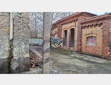Verlassener Industriebau aus gelben und roten Ziegeln mit bogenförmigen Fenster- und Türöffnungen, teils vergittert und mit Graffiti besprüht. Davor liegt ein rissiger, moosiger Betonhof mit einem Betonpfosten, einer bröckelnden Wand und kahlen Bäumen im Hintergrund.