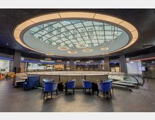 Moderne Innenansicht eines Gebäudes mit einem kreisförmigen Glasdach, das Tageslicht einlässt. Der Raum verfügt über Sitzbereiche mit blauen Sesseln, eine Rolltreppe und Beschilderungen für ein Restaurant und eine Bar.