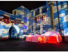 Nachtszene eines Innenhofs mit einer historischen Backsteinfassade, auf die bunte Lichtprojektionen mit Wolken, einem Flugzeug und einem Turmstrahler geworfen werden. Im Vordergrund stehen leuchtende, weiße Zeltpavillons mit roter Innenbeleuchtung.