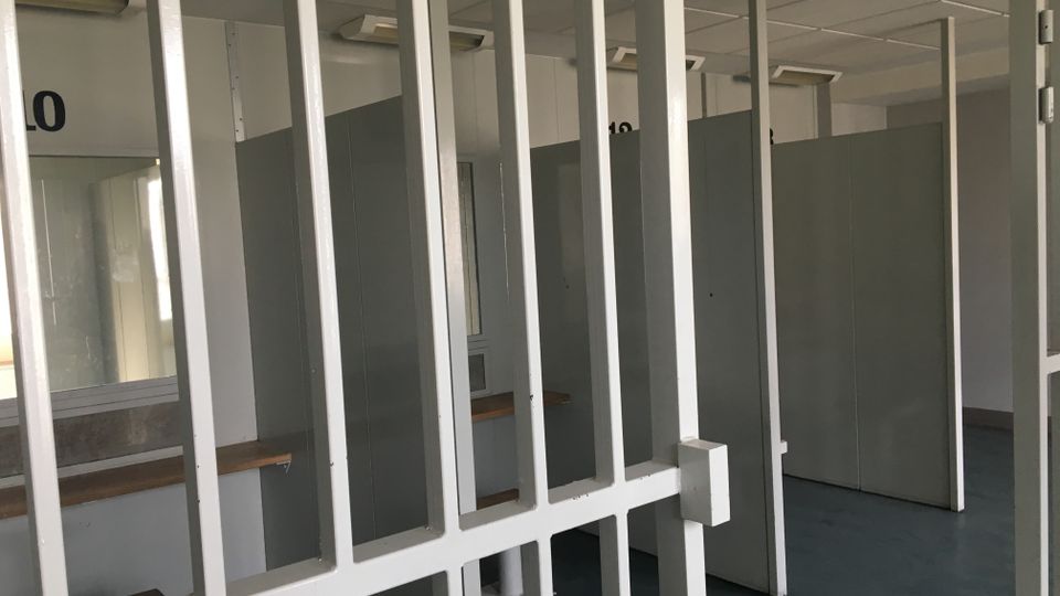 Blick durch ein weißes Metallgitter in einen Innenraum mit grauen Trennwänden und mehreren nummerierten Kabinen, beleuchtet von Deckenleuchten. Links sind ein Fenster und eine Bank zu sehen, der Boden ist dunkelblau.