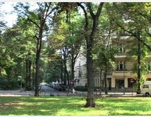 Grüne Parklandschaft mit hohen Bäumen, die eine Straße säumen; im Hintergrund sind elegante, helle Wohn- und Geschäftsgebäude zu sehen. Eine ruhig gelegene Stadtstraße führt durch die parkähnliche Umgebung.