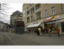 Straßenszene mit mehrstöckigen Wohn- und Geschäftsgebäuden, darunter ein Bäcker mit der Aufschrift „Bäcker Wiedemann“ und weitere Geschäfte. Im Vordergrund befindet sich ein Eingang zur U-Bahn-Station „Theodor-Heuss-Platz“ mit Glaskonstruktion und einem Werbeschild.