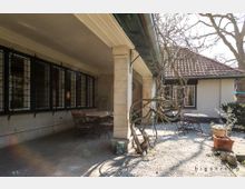 Überdachte Terrasse eines Hauses mit rustikalen Elementen, darunter eine Natursteinwand, schmiedeeiserne Gitter vor den Fenstern und ein geschwungener Baum vor der Terrasse. Im Außenbereich stehen Gartenmöbel auf einem kiesbedeckten Boden.