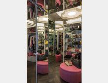 Ein begehbarer Kleiderschrank mit mehreren Spiegeltüren, gefüllten Regalen mit Handtaschen und Accessoires sowie Kleiderstangen mit Kleidung. Im Zentrum steht ein runder, pinker Hocker, und die Beleuchtung wird durch runde Deckenleuchten ergänzt.