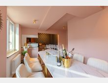 Ein kleiner Raum mit rosa Wänden und einer Küchenzeile im Hintergrund. Im Vordergrund steht ein langer Esstisch mit heller Marmoroptik, umgeben von gepolsterten weißen Stühlen, bestückt mit Gläsern, Champagnerflaschen und einem goldenen Kühler.