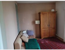 Kleines Wohnzimmer mit beiger Couch, mehreren Kissen, einem großen Holzschrank und einem Stehlampenständer. Auf dem Boden liegt ein dekorativer roter Teppich, und eine Wand ist mit Glasbausteinen gestaltet.