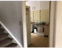 Blick in eine kleine Küche mit cremefarbenen Schränken, einem Herd mit Backofen und einer Spüle. Die Wände sind mit gelblichen Fliesen bedeckt, die mit farbigen Blumenmustern verziert sind, daneben befindet sich eine weiße Wand mit einer Uhr. Im Vordergrund ist eine Treppe erkennbar.