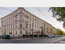 Straßenszene mit einem historischen Gebäude im Stil des 19. Jahrhunderts, das als Hotel genutzt wird. Das Gebäude hat eine helle Fassade mit dekorativen Fensterrahmen und liegt an einer von Straßenbahnschienen und Bäumen gesäumten Straße.