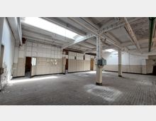 Großer, leerstehender Industrieraum mit abgenutztem Holzboden, weißen Wänden und einer Decke mit sichtbaren Trägern sowie mehreren Oberlichtern. An den Seiten befinden sich elektrische Leitungen und ein veralteter Schaltschrank, die Raumaufteilung wird durch brüchige Trennwände und offene Türen ergänzt.