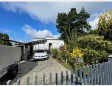 Moderne Wohnhausfassade mit weißem Putz, davor eine gepflasterte Einfahrt mit einem geparkten Auto unter einem gebogenen Carport-Dach. Rechts ein Garten mit blühenden Sträuchern und Bäumen, im Hintergrund blauer Himmel mit Wolken.
