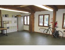 Ein schlichter multifunktionaler Raum mit Holzelementen, sichtbaren Deckenbalken und großen Fenstern, die Tageslicht hereinlassen. An der Wand stehen ein weißes Regal mit Fächern, ein Metallschrank und ein Fahrrad; zusätzlich sind ein Feuerlöscher und ein Heizgerät angebracht.