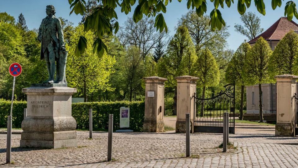 Cobblestone plaza at the entrance to a landscaped park, featuring a bronze statue on a stone pedestal and an ornate wrought-iron gate between stone pillars. Trees and trimmed hedges line the background, with a light-colored building topped by a red-tiled roof visible beyond the gate.
