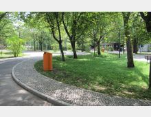 Ein Park mit gepflasterten Wegen, Grünflächen und schattenspendenden Bäumen. Im Vordergrund steht ein orangefarbener Abfalleimer, im Hintergrund sind Bänke und eine kleine Holzhütte zu erkennen.