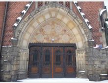 Großes gotisches Kirchenportal aus Holz und Metall mit aufwendigen Schnitzereien und dekorativen Eisengittern, umgeben von einem spitzbogigen Sandsteinrahmen mit Verzierungen. Darüber befindet sich ein farbig gestaltetes, florales Mosaik sowie Backsteinmauerwerk; der Boden ist schneebedeckt.