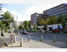 Straßenszene an der Fehrbelliner Straße mit einer mehrspurigen Fahrbahn, beidseitigem Gehsteig und parkenden Autos. Im Hintergrund befinden sich ein moderner Ziegelbau, ein traditionelles Wohngebäude und Bäume entlang der Straße.