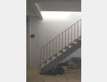 Eine minimalistische Treppe mit Metallgeländer und Holzstufen vor einer weißen Wand. Im unteren Bereich befinden sich gestapelte Bücher und lose Gegenstände auf einem Holzboden.