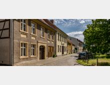 Pflastersteinstraße mit historischen Wohnhäusern in einer ruhigen Altstadt. Links sind mehrstöckige Gebäude mit verputzten und farbigen Fassaden, rechts ein Baum und parkende Autos.