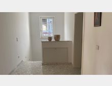 Heller Flur mit Fenster am Ende, gefliesten Marmorboden und einer halbhohen Ablage mit zwei Dekovasen. Rechts ist eine Zimmertür und ein an der Wand hängendes Bild erkennbar.