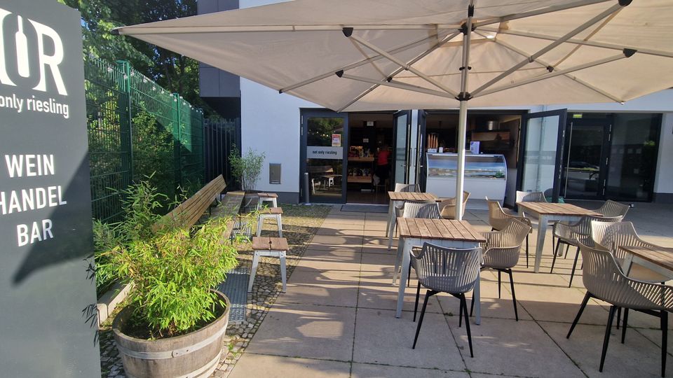 Außenbereich einer Weinbar mit Holz-Tischen und Kunststoffstühlen unter großen weißen Sonnenschirmen vor einem modernen Gebäude mit geöffneten Glastüren. Links steht ein Schild „NÖR – not only riesling – Wein Handel Bar“, daneben Holzbank, Zaun und Pflanzkübel.