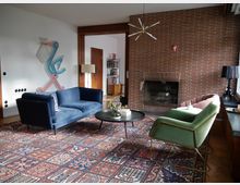 Wohnzimmer mit einer gemusterten Teppichfläche, einem blauen Sofa, einem rosa Sessel und einem grünen Sessel. Im Raum befinden sich ein gemauerter Kamin vor einer Ziegelwand, ein runder Couchtisch sowie Wanddekor in Form eines bunten Vogels.