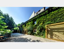 Ein mit Efeu bewachsenes Gebäude mit großen gelben Fenster- und Türrahmen in einem gepflasterten Hof, umgeben von Bäumen und Sträuchern unter einem klaren blauen Himmel.