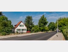 Wohnstraße mit einem kleinen weißen Einfamilienhaus mit rotem Satteldach und grünen Fensterläden, umgeben von Bäumen und einem Zaun. Rechts führt eine schmale asphaltierte Straße mit Gehweg und Straßenlaterne entlang.