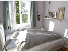 Schlafzimmer mit einem großen Bett, grauer Decke und gestreiftem Kissen, das von Tageslicht durch ein großes Fenster mit weißen Vorhängen erleuchtet wird. An der Wand hängt ein weißes Regal mit gerahmten Bildern und Dekorationen, und ein Heizkörper befindet sich unter dem Fenster.