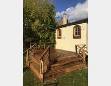 Ein kleiner Holzbau mit cremefarbener Fassade und zwei Fenstern mit Holzrahmen, daneben eine große Holzterrasse mit Geländer. Im Hintergrund sind ein Baum und ein gemauertes Gebäude sichtbar.